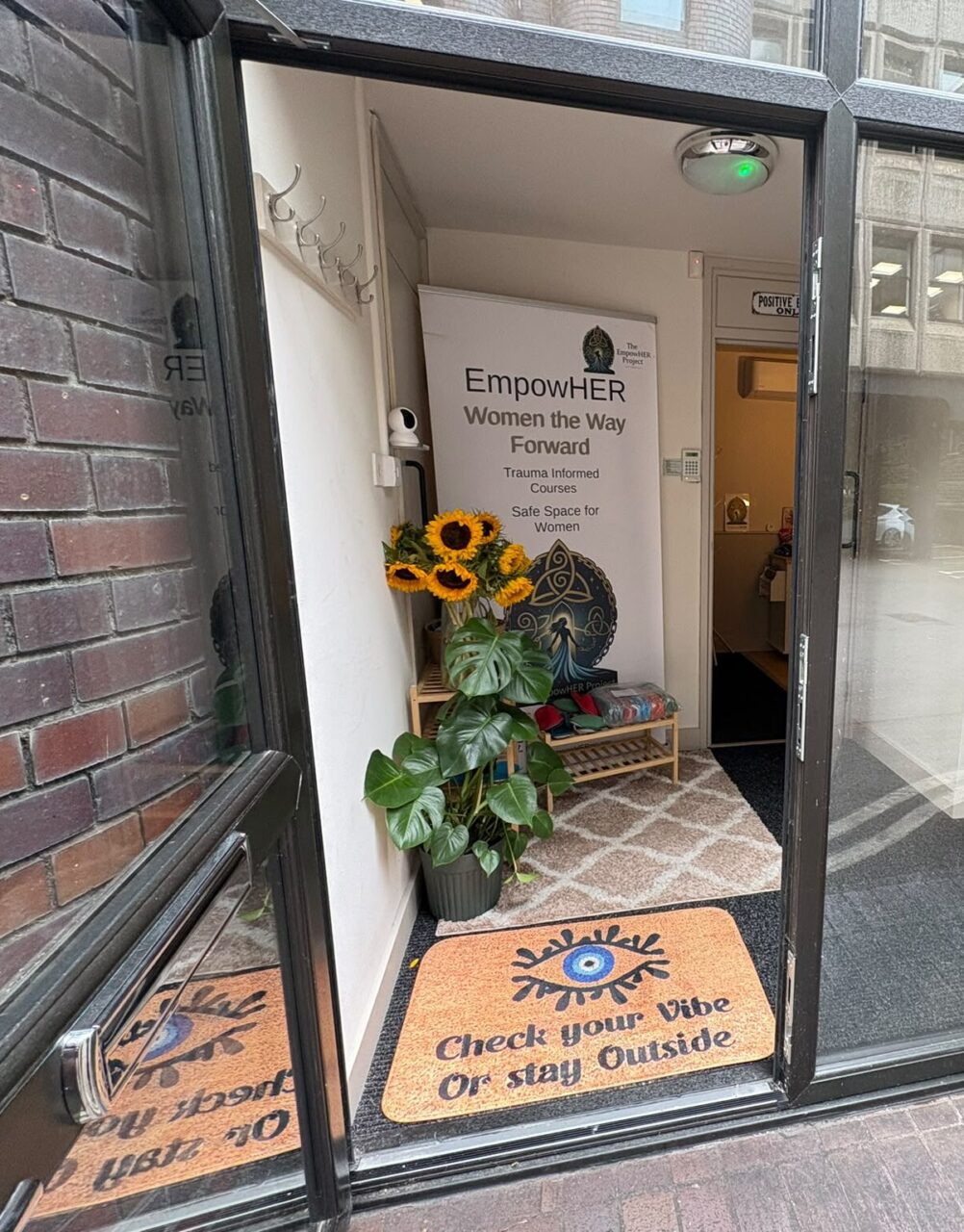 Entrance to the EmpowHER Project centre, showing an open glass doorway, a welcome mat reading “Check your Vibe or stay Outside”, potted plants with sunflowers, and a sign promoting trauma-informed courses and a safe space for women.