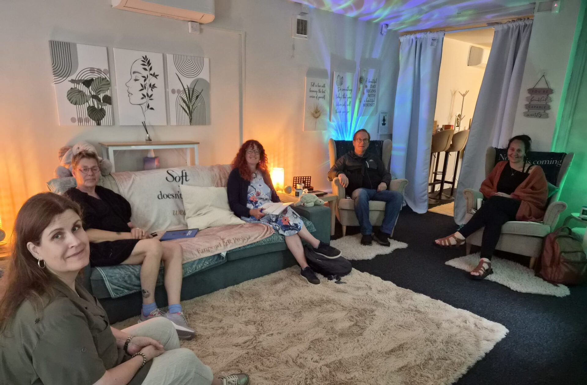 Five visitors seated in a comfortable wellbeing room at the EmpowHER centre. The space features sofas, armchairs, soft furnishings, calming lighting effects and wellbeing-themed wall artwork.